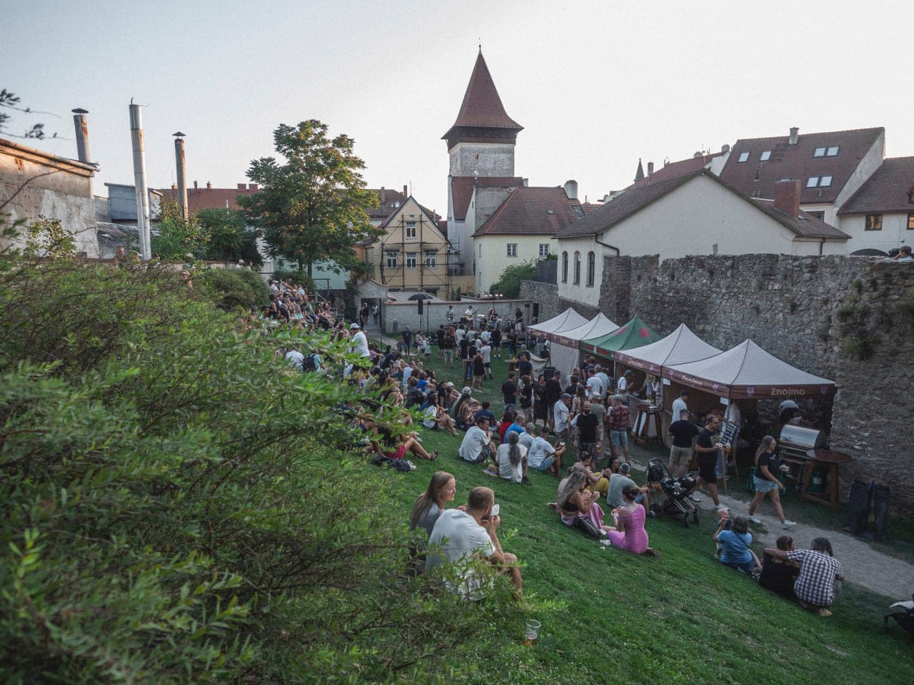 Lidé sedí na travnatém valu pod hradbami, dole stánky a pódium, v pozadí věž