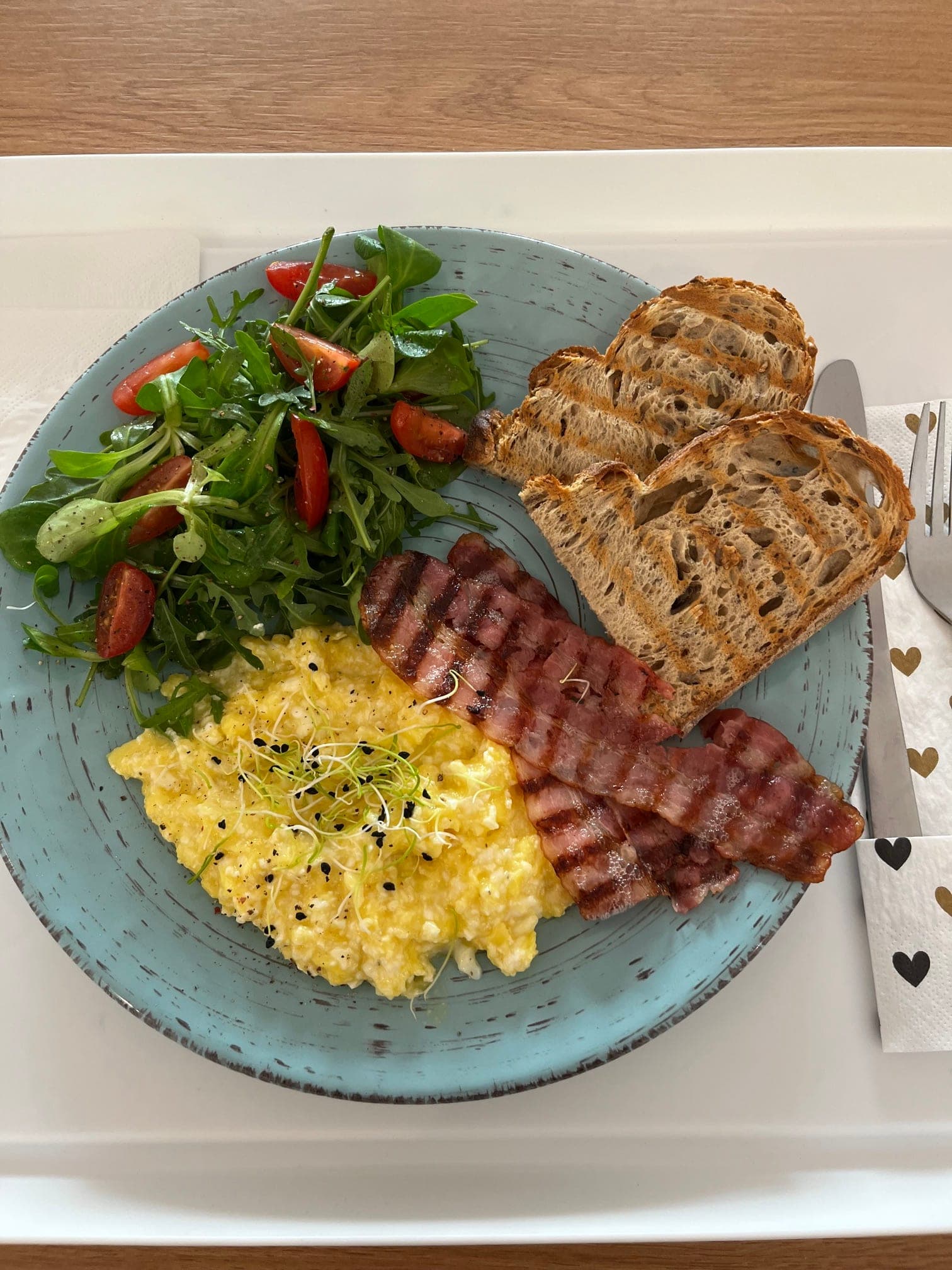 Rührei mit gegrilltem Speck, geröstetem Brot und Rucola-Tomaten-Salat auf einem türkisfarbenen Teller