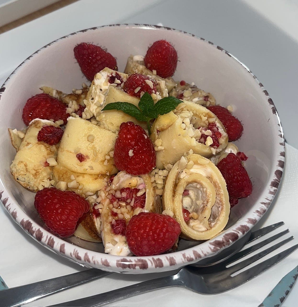 Eine Schale gefüllter Palatschinkenröllchen mit Himbeeren, gehackten Nüssen und einem Minzblatt
